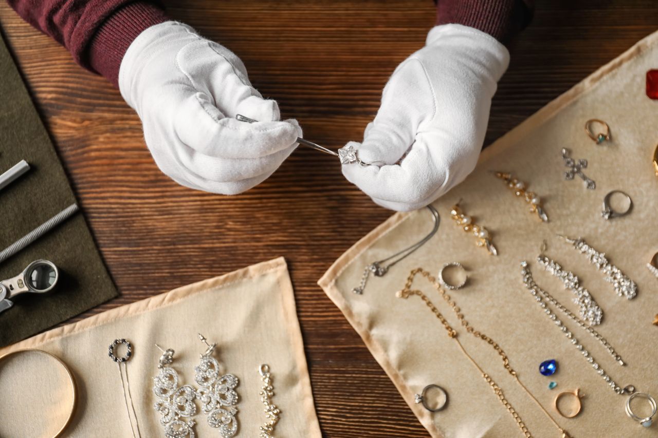 A view of a jeweler wearing white gloves as they inspect a diamond ring, surrounded by elegant jewelry on soft fabric.