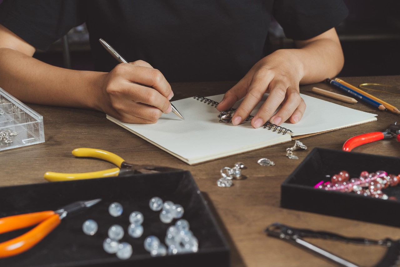 A close-up of an artisan sketching jewelry designs, surrounded by tools, beads, and gemstones atop a wooden desk.