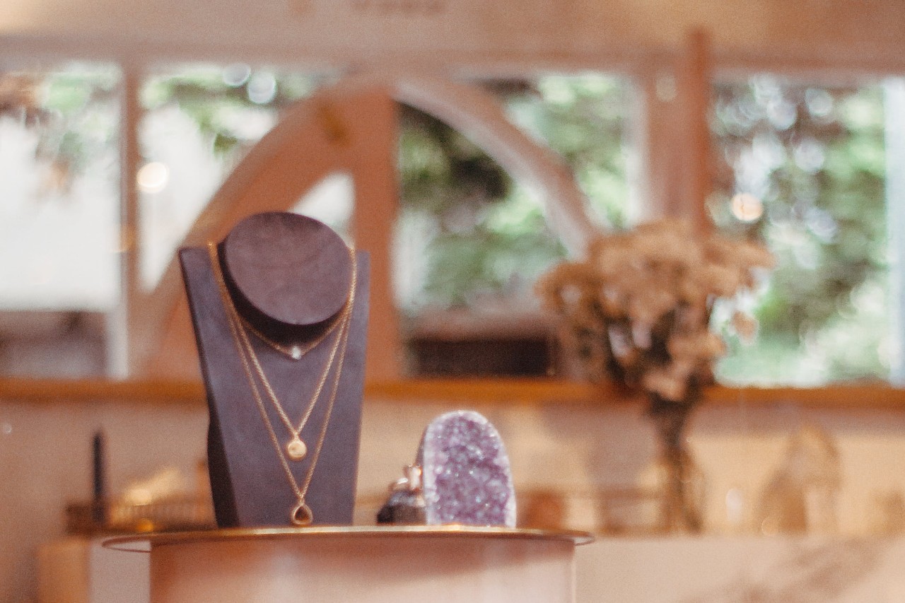 A layered gold necklace is displayed on a black jewelry stand beside a purple crystal inside a softly lit boutique with blurred greenery in the background.