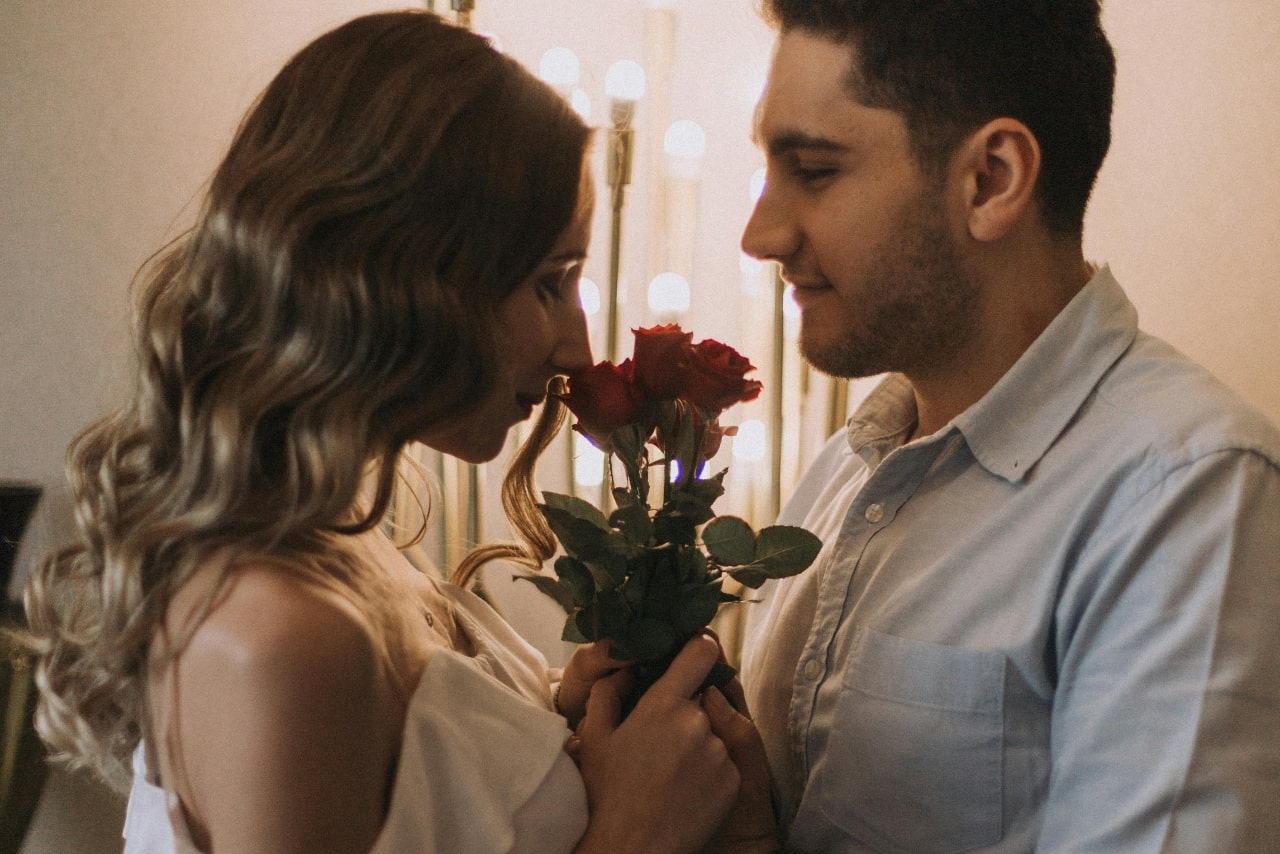 A man and woman standing very close and facing each other, the woman smelling red roses with romantic lighting.