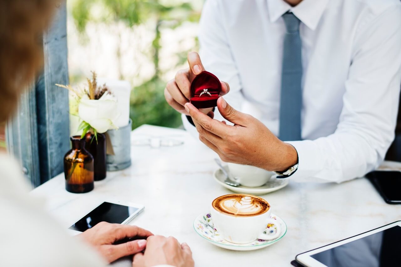 A man in a white shirt and grey tie proposes with an open red ring box with an engagement ring across a café table during a proposal