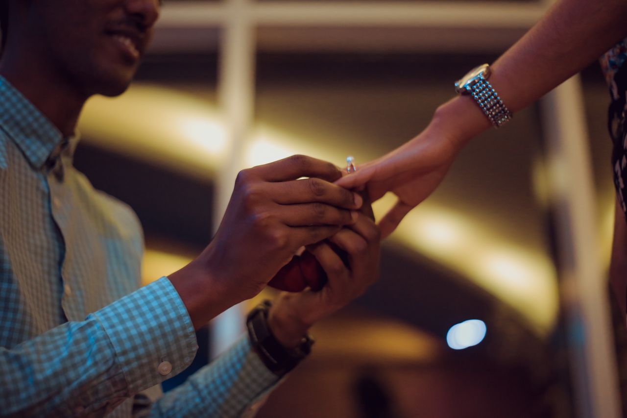 A man places an engagement ring on a woman&rsquo;s finger while holding a red ring box in a warmly lit setting