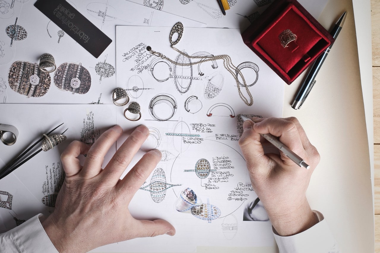 Hands sketch and annotate jewelry designs on paper, surrounded by rings, a necklace, tools, and a red ring box on a designer&rsquo;s worktable