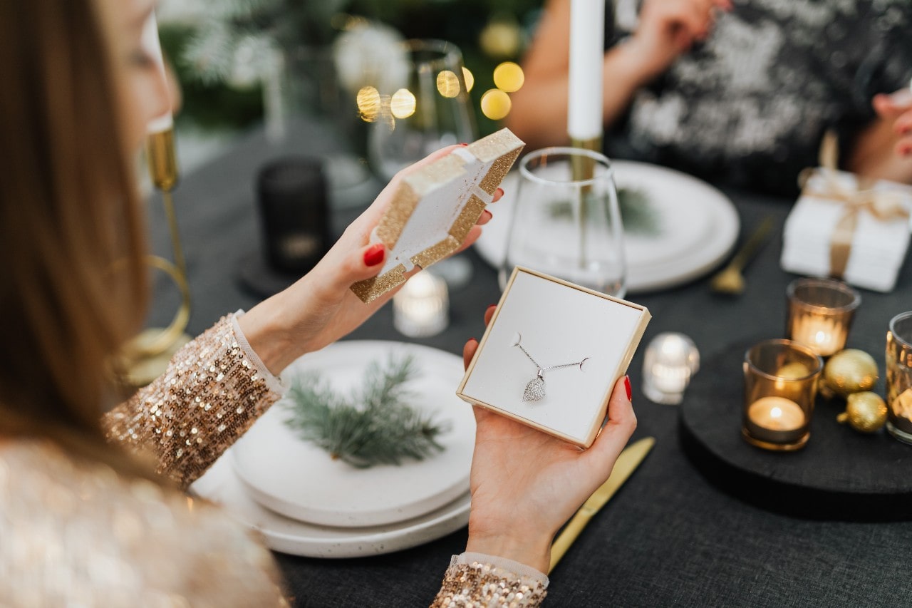 A woman opens a gift box to reveal a delicate diamond necklace at a festive, candlelit dinner table decorated with holiday accents