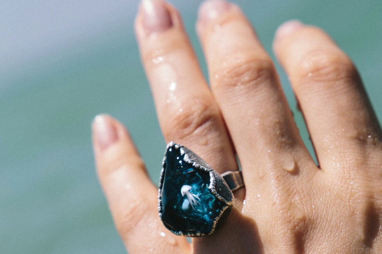 A hand wearing a large, teardrop-shaped blue ring with a silver band is held up, water droplets on the fingers.