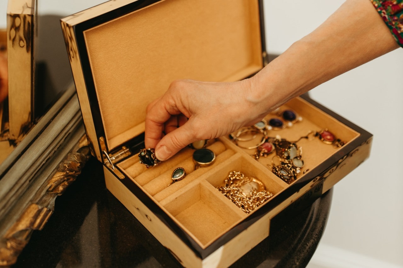 A hand selecting a ring from an open jewelry box filled with assorted gemstone rings, bracelets, and chains.