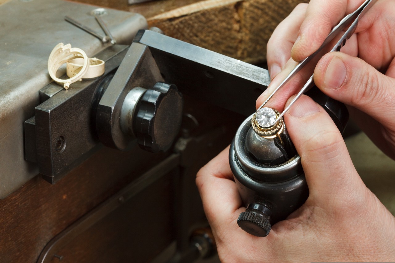 Close-up of a jeweler's hands using tweezers to attach a large gem to a gold ring.