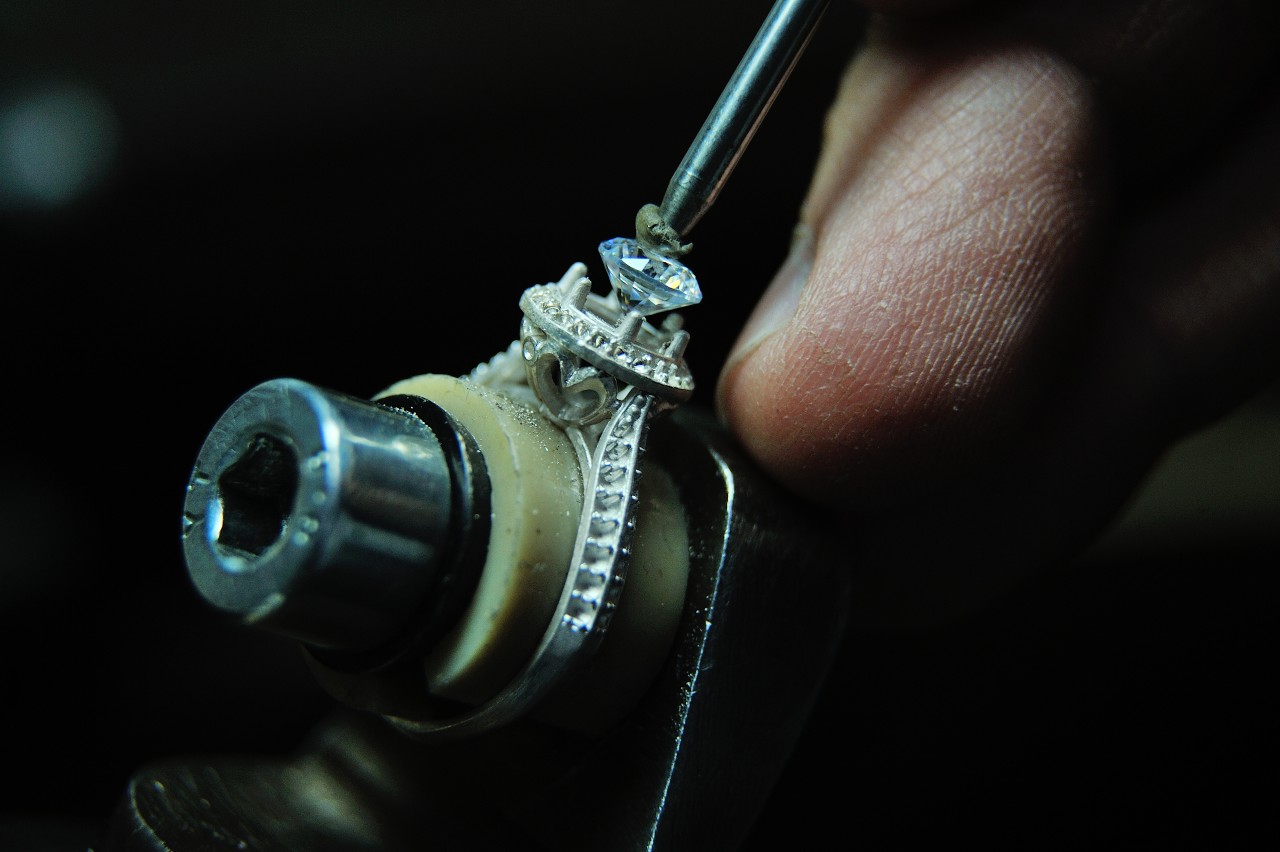 Close-up of a jeweler&rsquo;s hand delicately working on a diamond ring with a small tool.