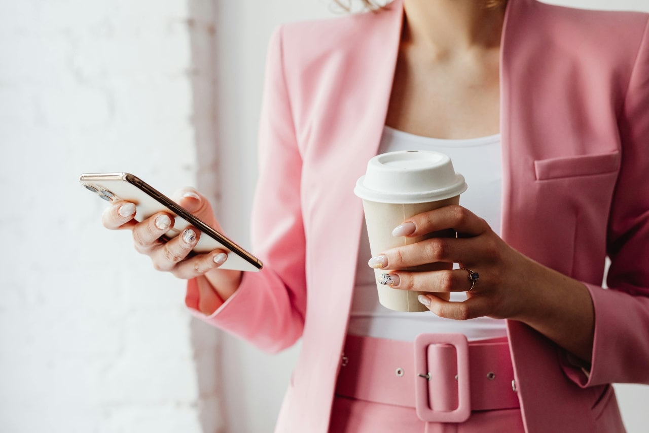 A woman in a pink suit, holding a coffee cup and her phone while she types a text out.