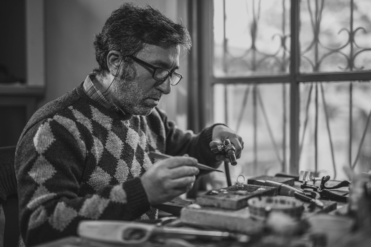 Black and white image of a jeweler in a patterned sweater, carefully working with tools at a table by a window.