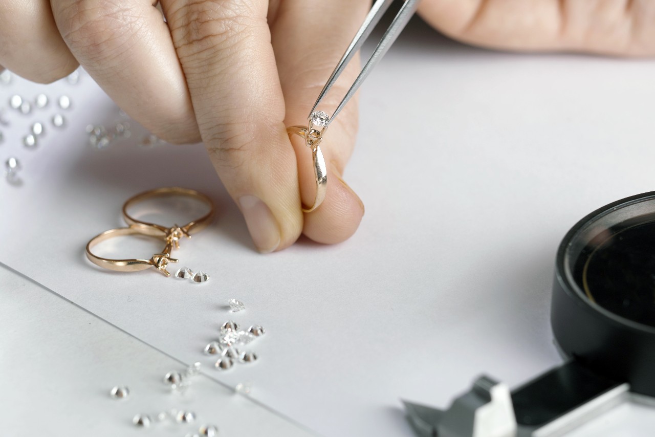 A jeweler sets a diamond in a gold ring by tweezers, surrounded by loose diamonds, another ring, a magnifying glass, and a caliper.