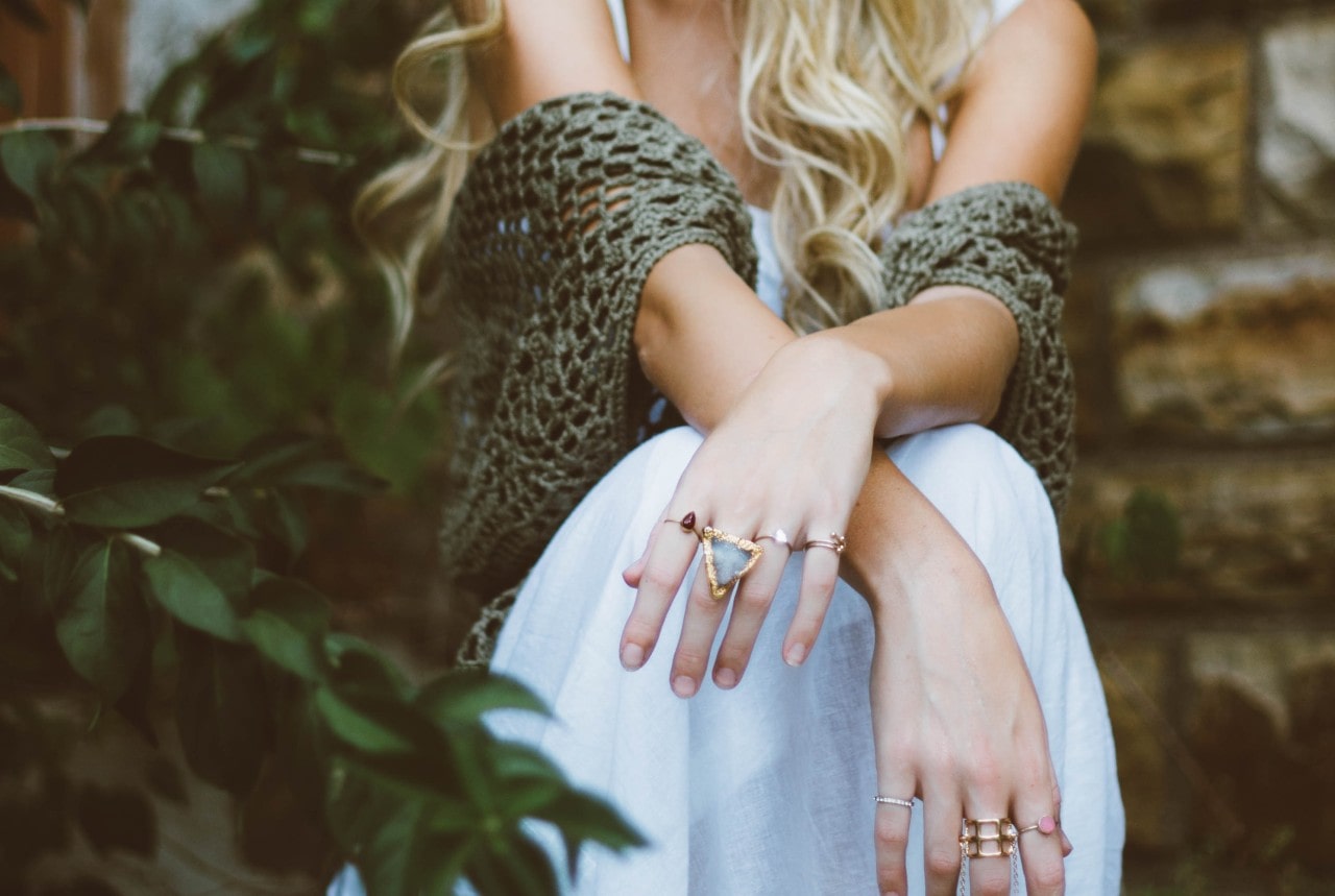 A woman in white dress and olive crocheted shawl sitting outdoors, showcasing multiple gold statement rings on both hands.