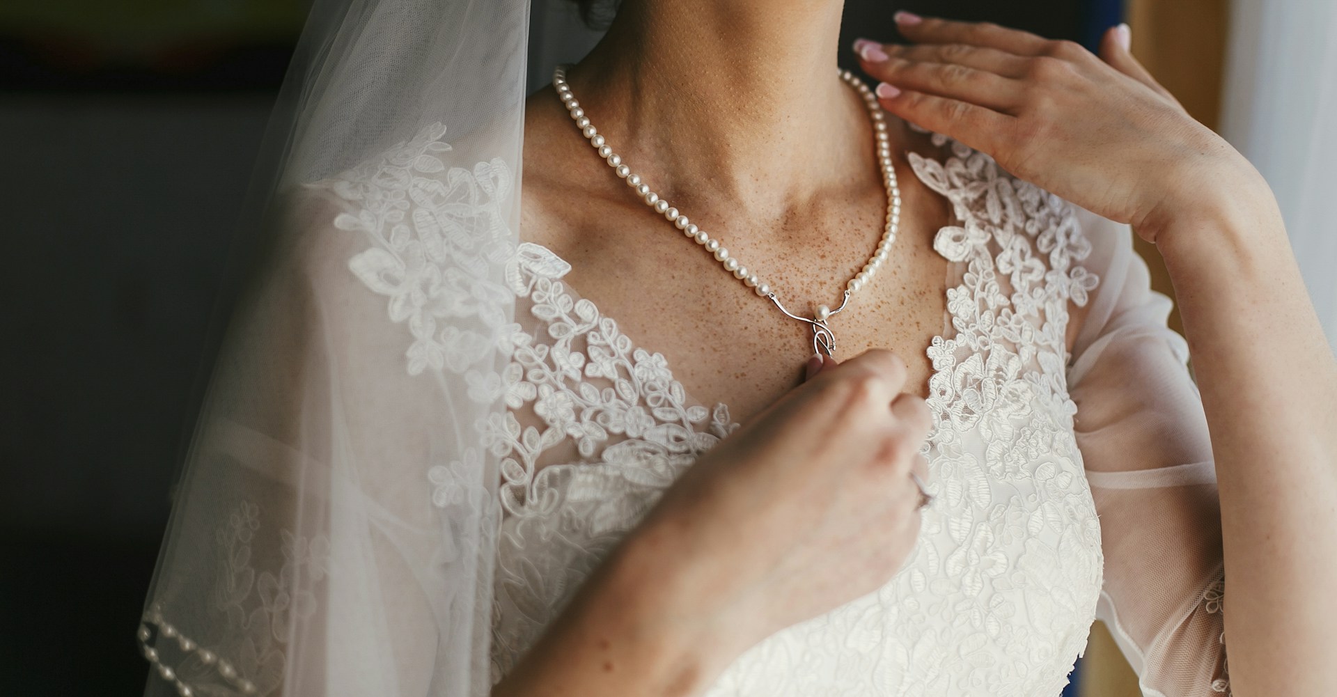 The bride in a white lace wedding dress gently adjusts a pearl necklace, her veil softly draping around her.