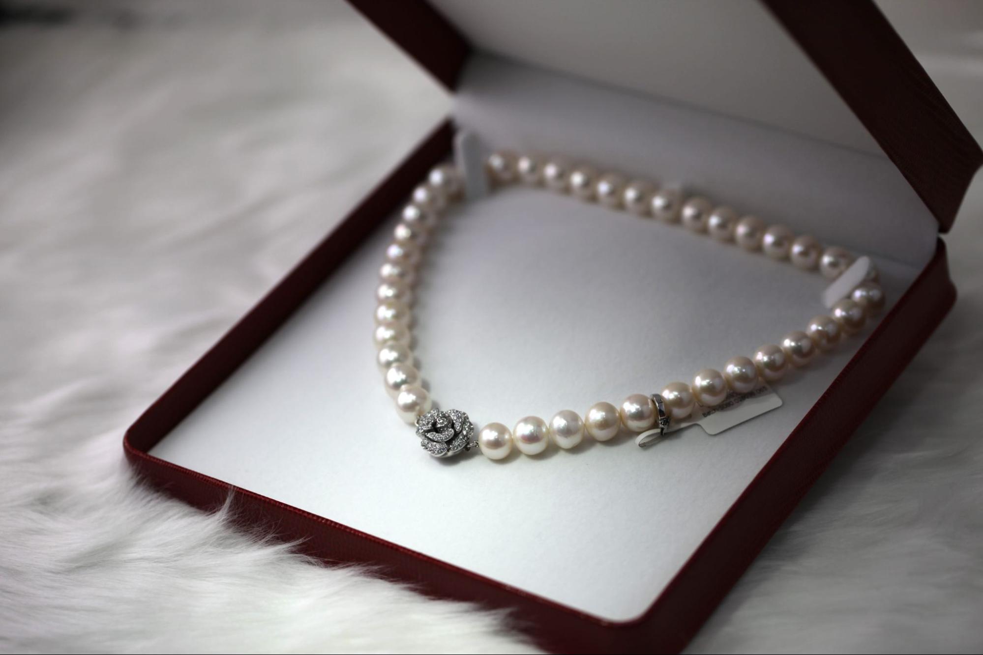 A pearl necklace with a flower-shaped clasp is elegantly displayed in an open jewelry box on a white fur surface.