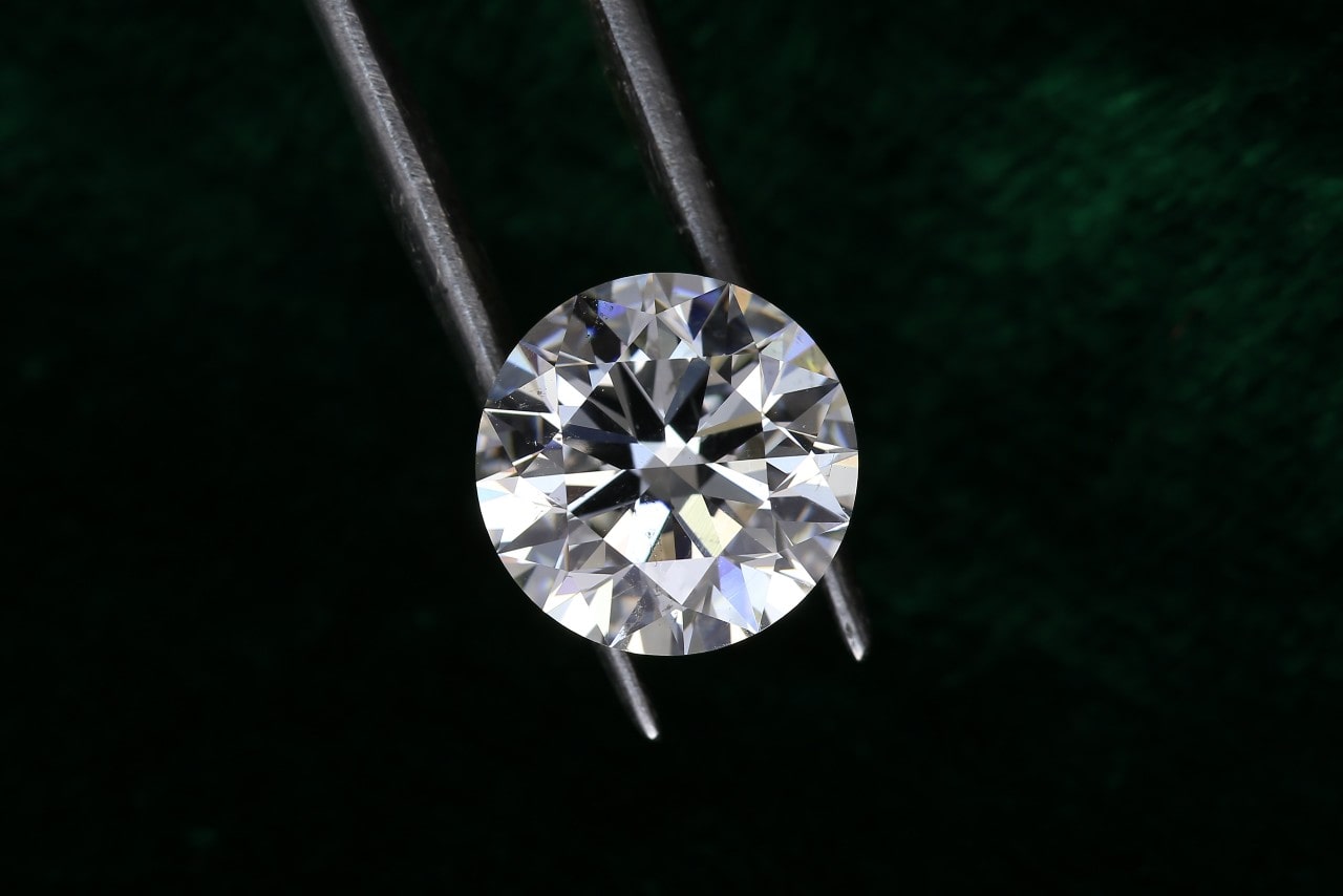 A close up of a round brilliant cut diamond held by tweezers against a dark green background