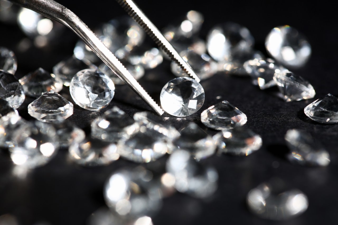 A dozen loose diamonds scattered all over black surface, with one being held by tweezers.