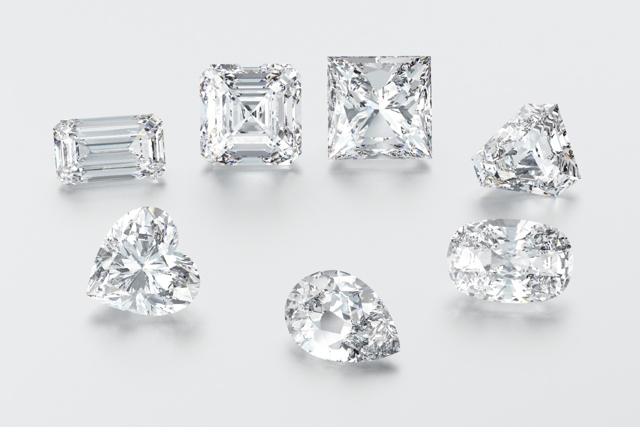 A close up of an emerald, cushion, heart, princess, oval, and pear shaped diamonds on a white surface.