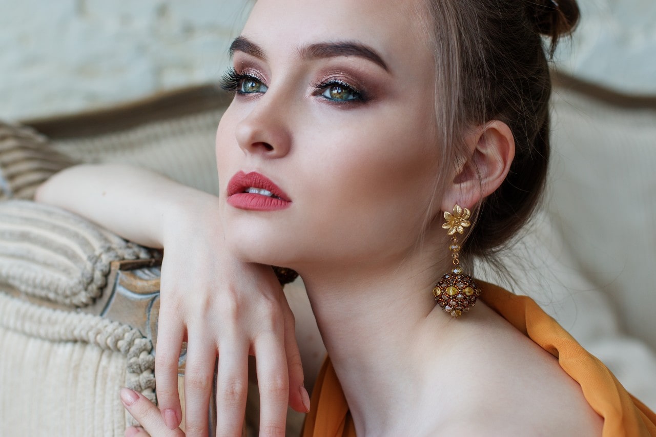 A woman with an elegant updo gazes thoughtfully, wearing sparkly eye makeup and intricate gold earrings.