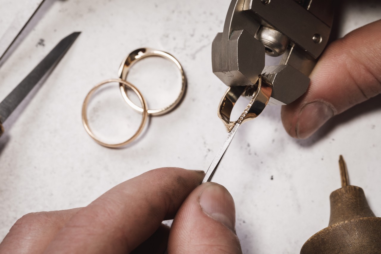 A close up of a jeweler repairing a yellow gold wedding band while two others lay on the white background.