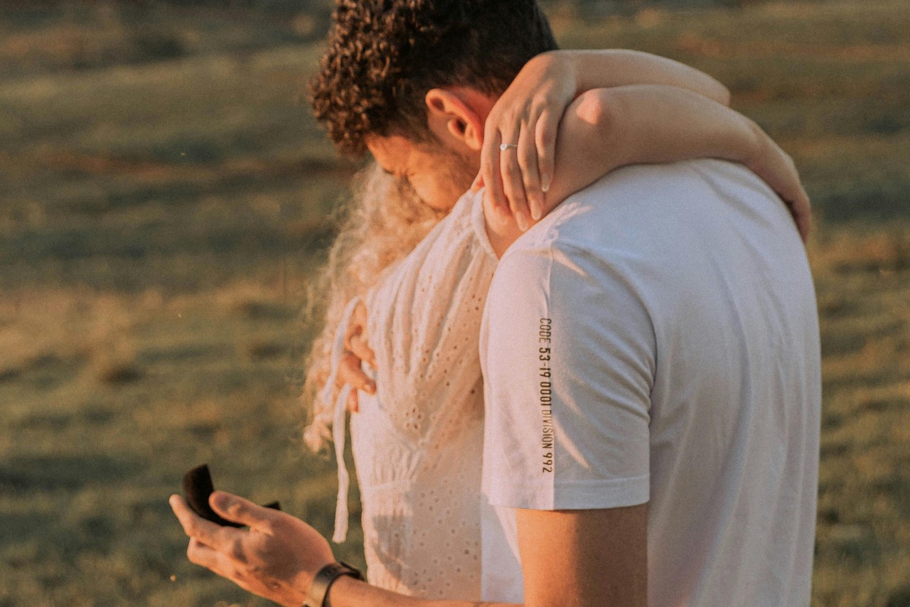 A woman wrapping her arms around a man’s neck in an open field, as he holds an open black ring box.