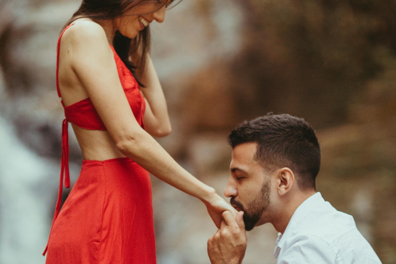 A man kneeling and kissing his partner’s hand during a proposal, with her wearing a red dress.