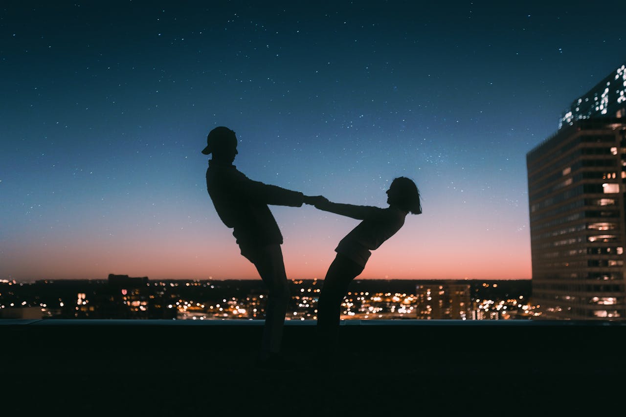 A backlit image of a couple dancing on a rooftop during dusk, the city skyline behind them.