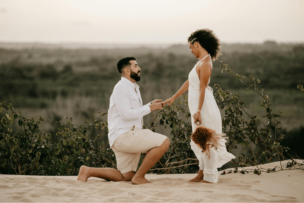 A man kneeling on sand while holding his partner’s hand as she holds a bouquet of flowers.