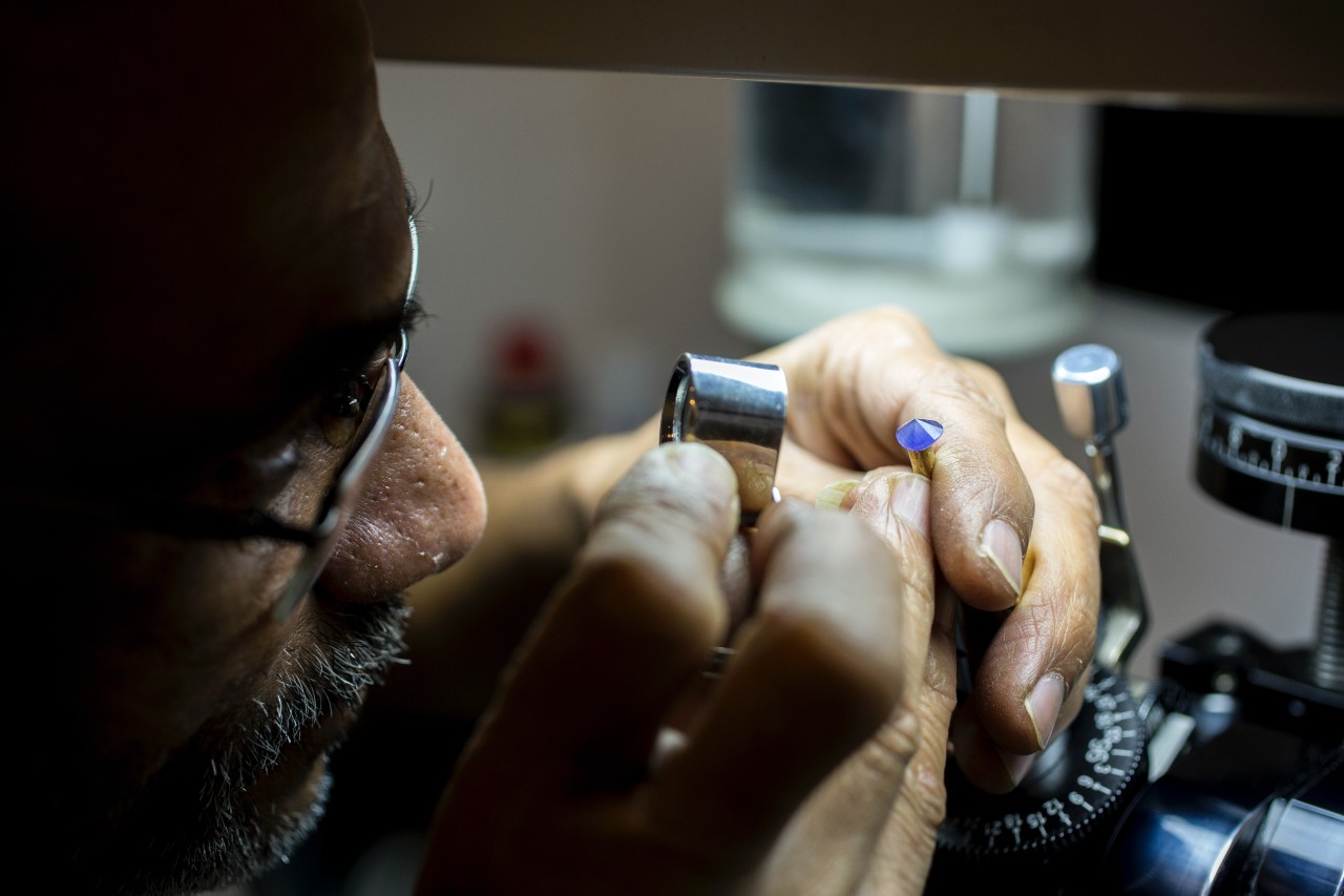 A jeweler closely examines a blue gemstone under magnification while holding it in a faceting machine.