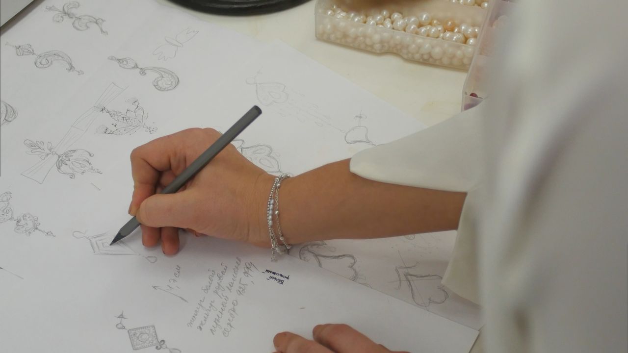 A designer sketches intricate jewelry designs on paper beside a tray of pearls.