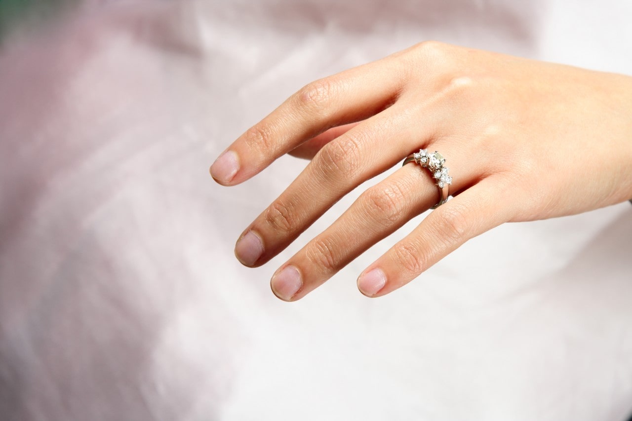 Close up of a hand adorned with a diamond engagement ring, against a soft white background.