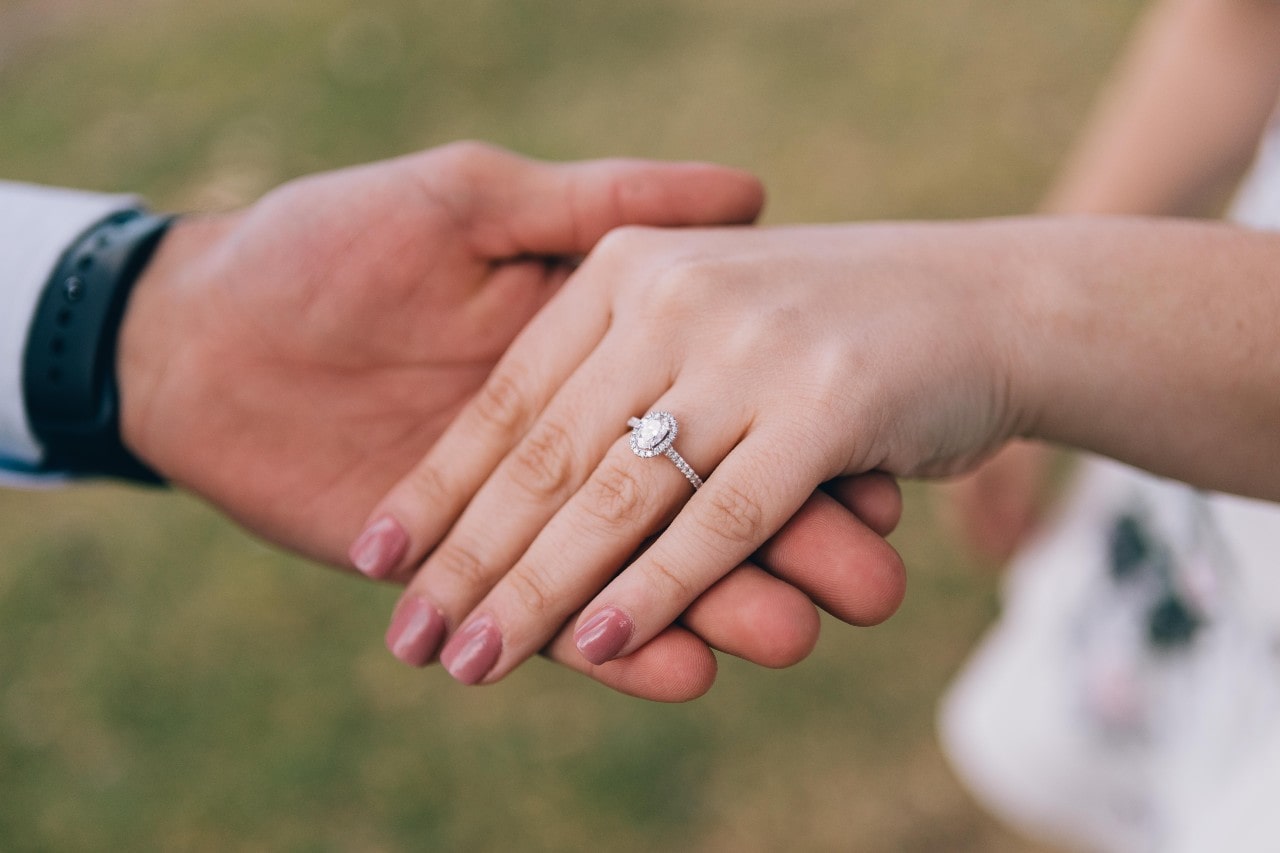 A close-up of a couple holding hands against a blurred green background, an elegant diamond halo engagement ring on the woman’s manicured hand.