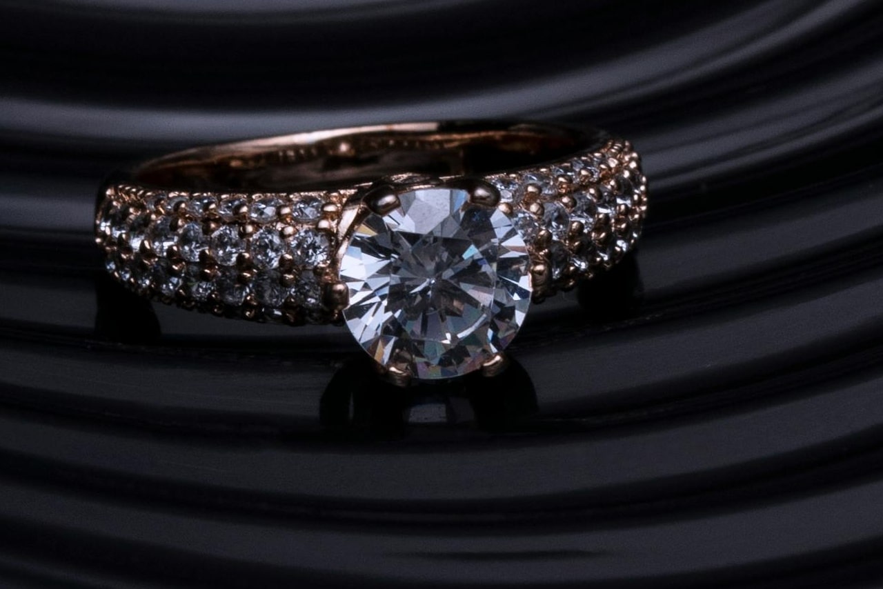 A close-up of a rose gold solitaire ring with pavé set diamonds along the band, set against a striped black background.