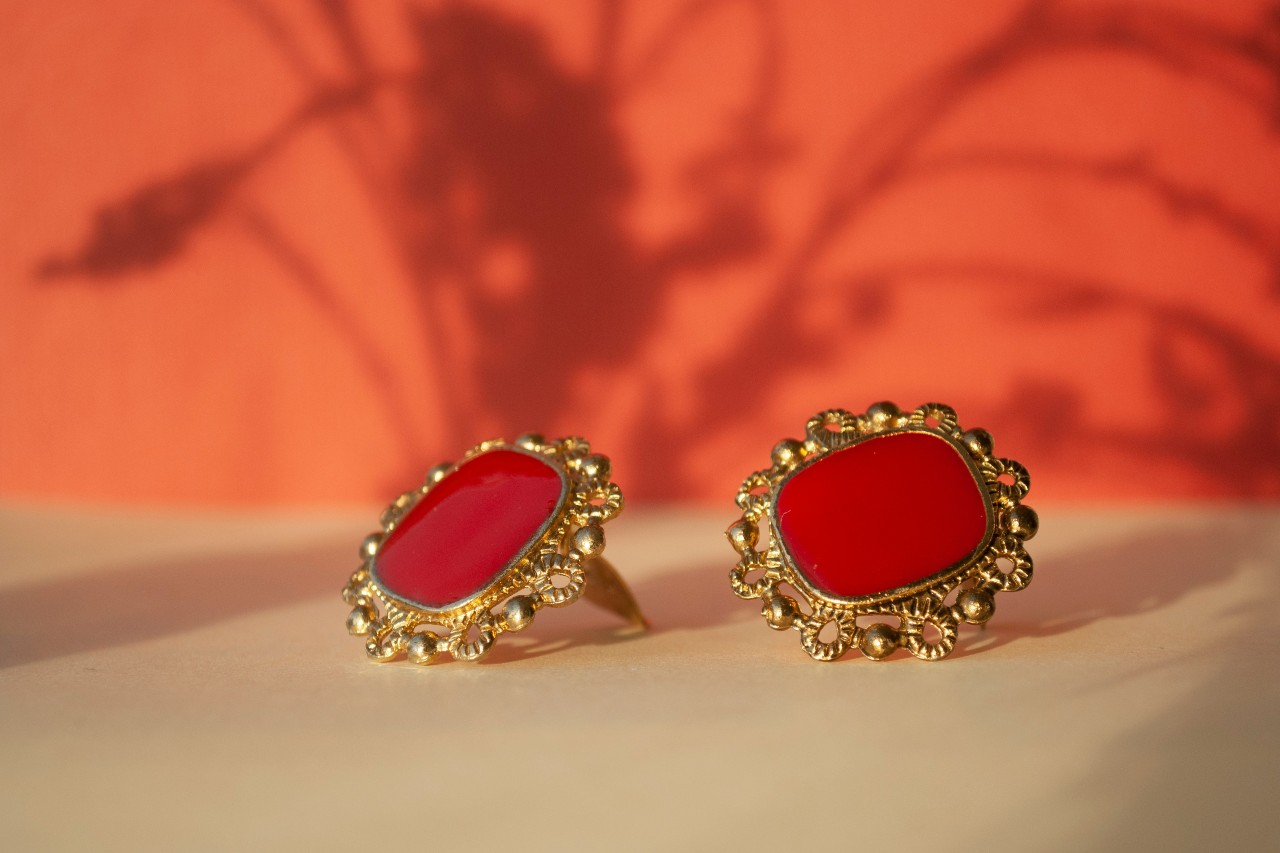 A close up of vintage yellow gold stud earrings with red center stone and intricate metalwork against an orange background. A close up of vintage yellow gold stud earrings with red center stone and intricate metalwork against an orange background.