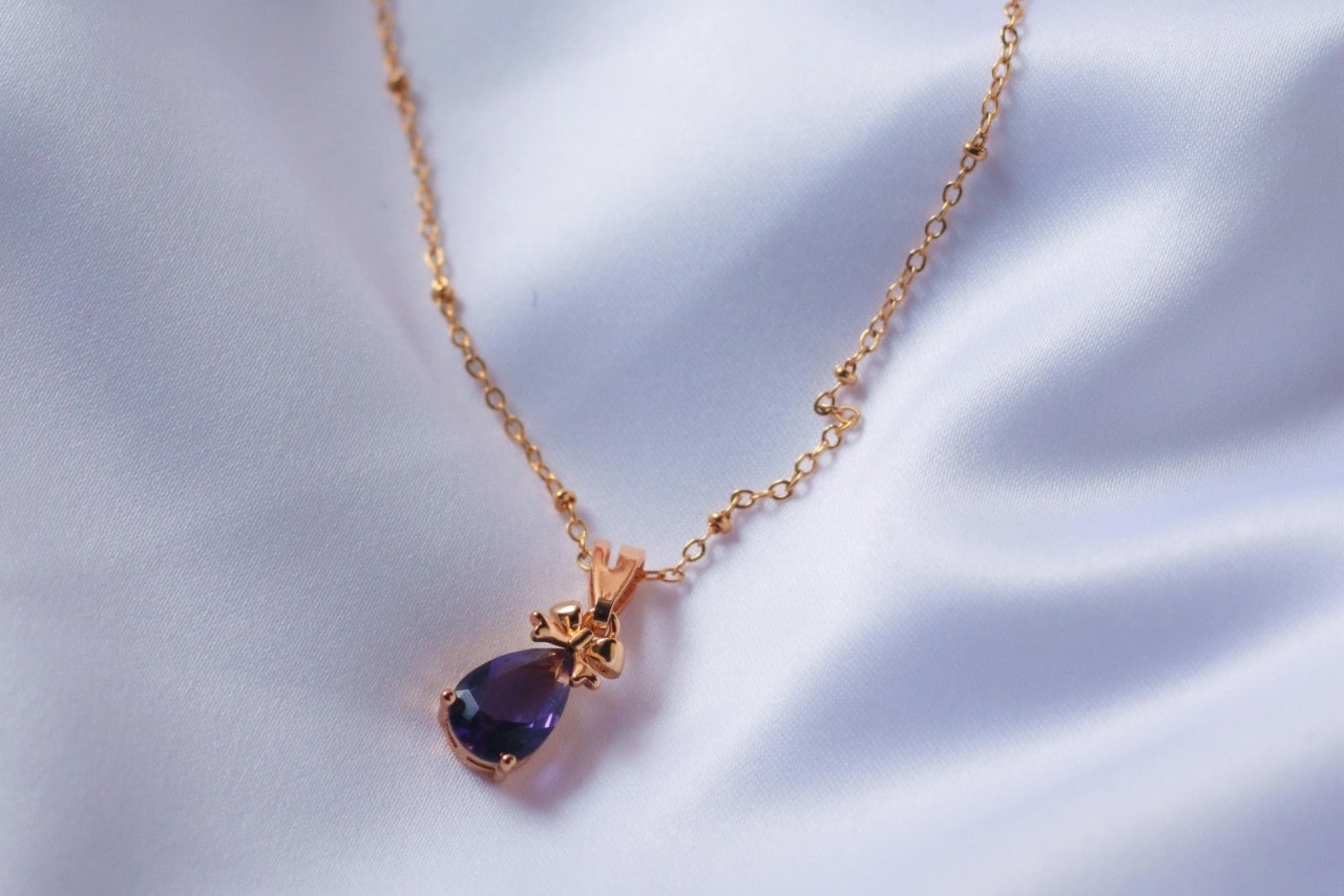 A yellow gold drop pendant with pear shaped purple gemstone against a white fabric. A yellow gold drop pendant with pear shaped purple gemstone against a white fabric.