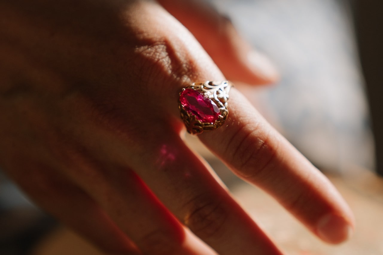 A close up of a vintage yellow gold ring with large ruby on a finger in a sunlight room A close up of a vintage yellow gold ring with large ruby on a finger in a sunlight room