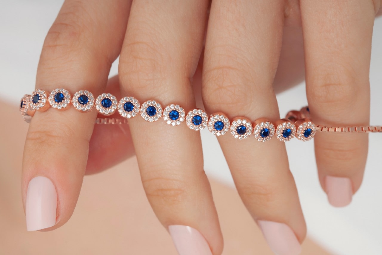 A rose gold halo sapphire tennis bracelet being held by hand with manicured nails. A rose gold halo sapphire tennis bracelet being held by hand with manicured nails.