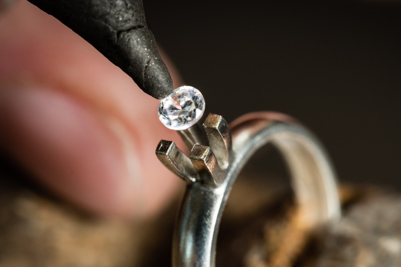 A close up of a jewelry expert working on placing a shiny diamond into its silver ring holder.