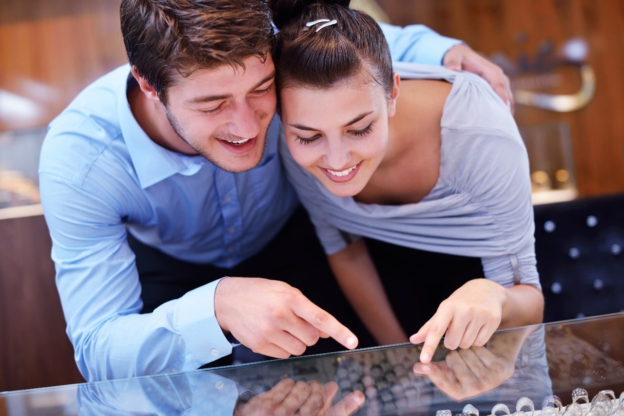An excited couple pointing and smiling at a jewelry case, with the man wrapping his arm around the woman.