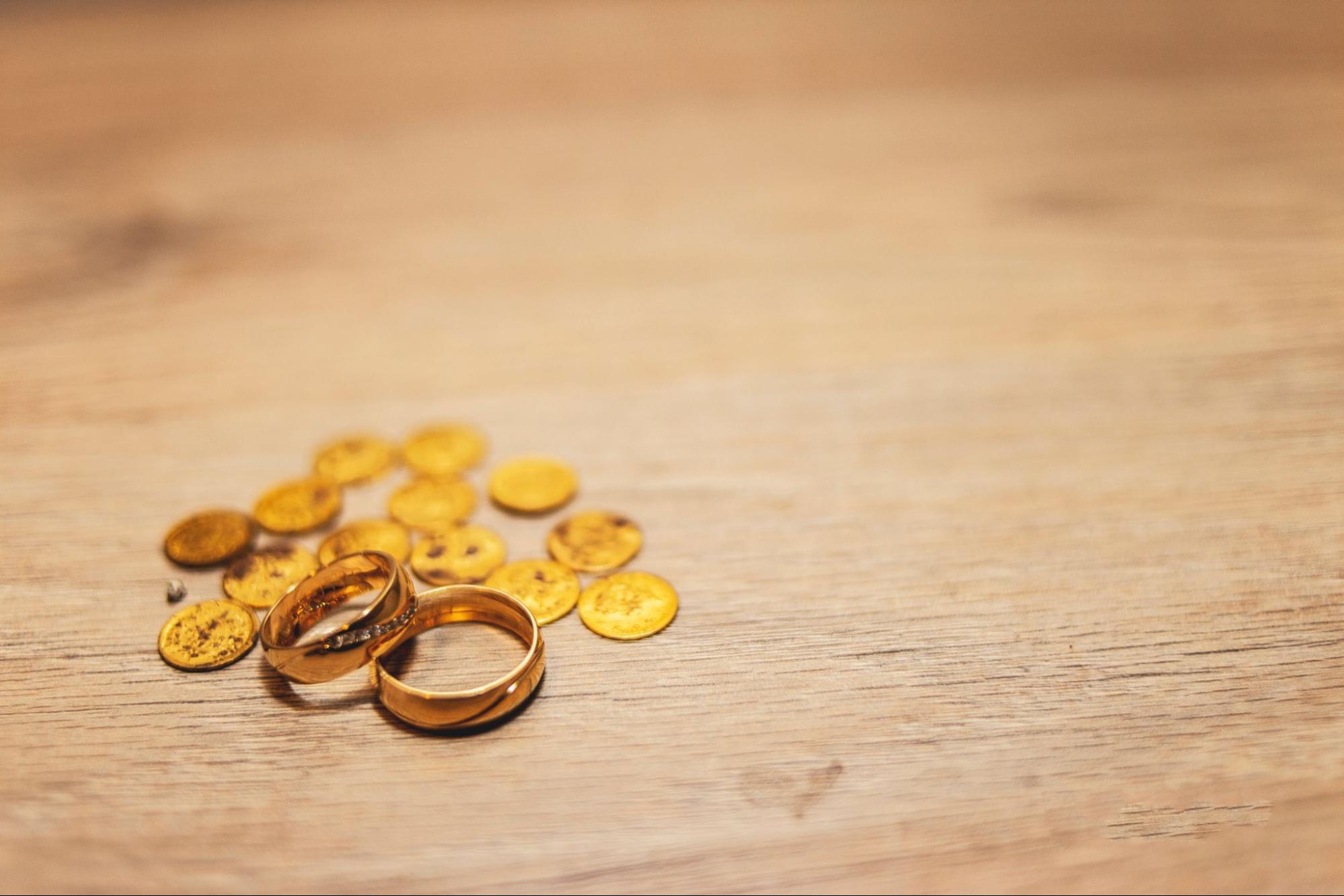 A close up of two yellow gold wedding bands and several gold coins on a wooden surface.
