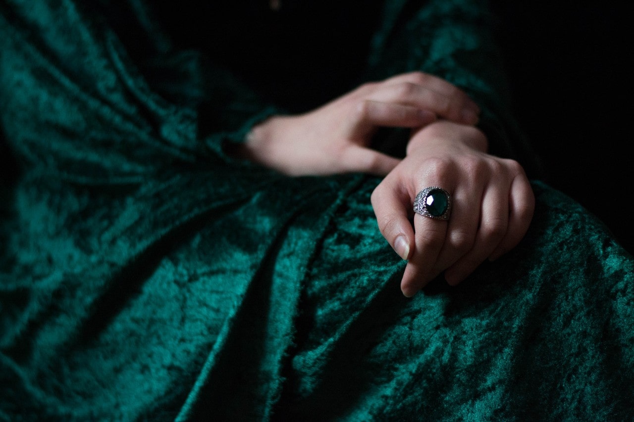 A close-up of a woman sitting in a deep green velvet dress with a massive emerald ring on her hand.